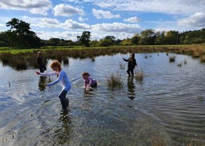 4 children playing in a large puddle extending into the distance. One of the children is crawling through the puddle in a green field with blue skies and scattered fluffy clouds above.