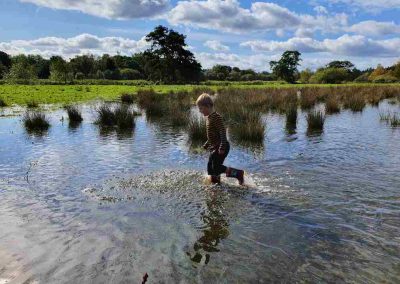 A large puddle of water extending into the distance with a child wading through it. The sky is blue with scattered fluffy clouds above a green field